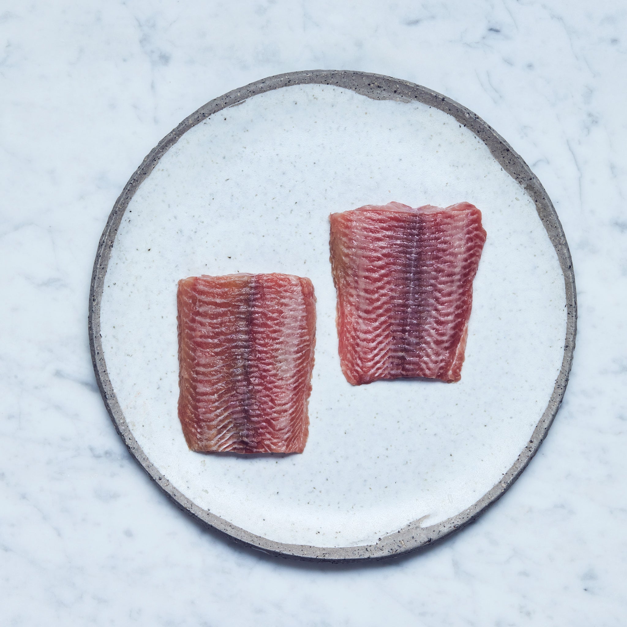 Ein runder Teller mit frischen Wels-Filet in zwei Stücke geschnitten auf einer Marmoroberfläche.