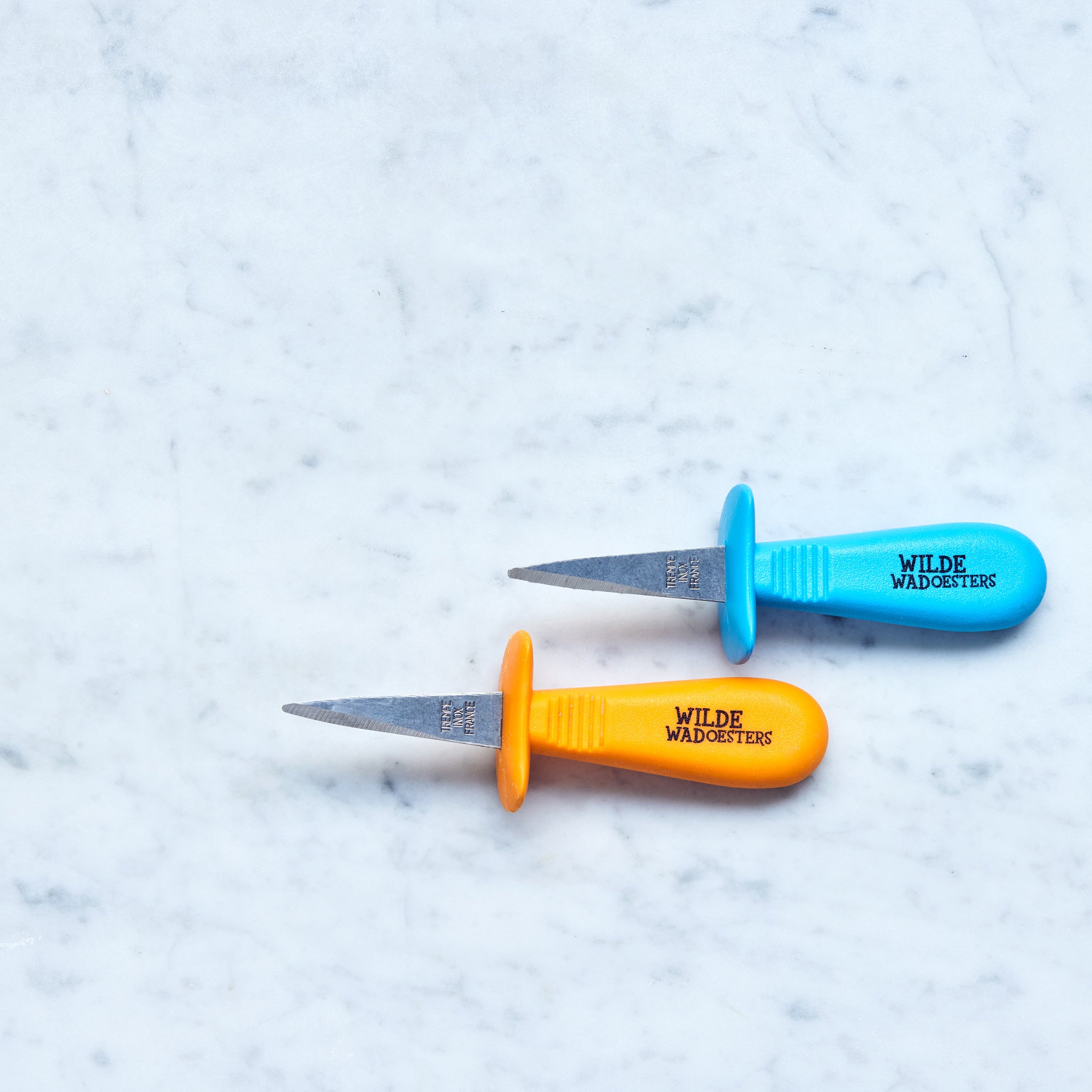 Zwei Austernmessern, blau und orange, auf einer Marmoroberfläche.