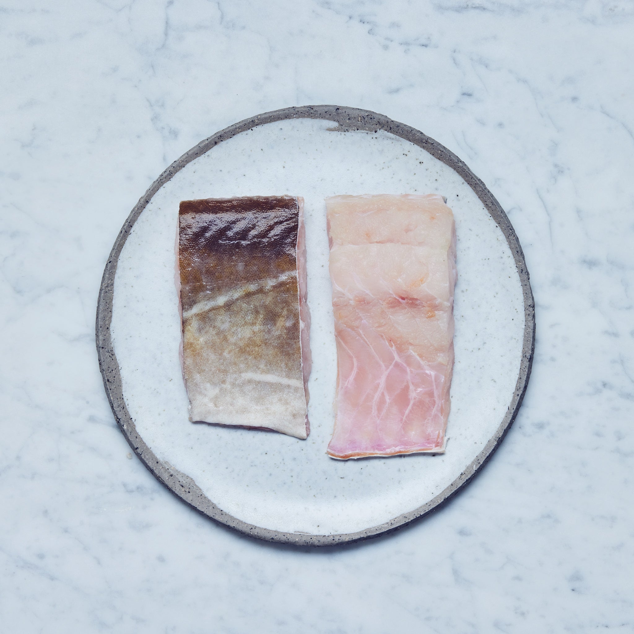 Ein runder Teller mit frischen Leng-Filet in zwei Stücke geschnitten auf einer Marmoroberfläche.