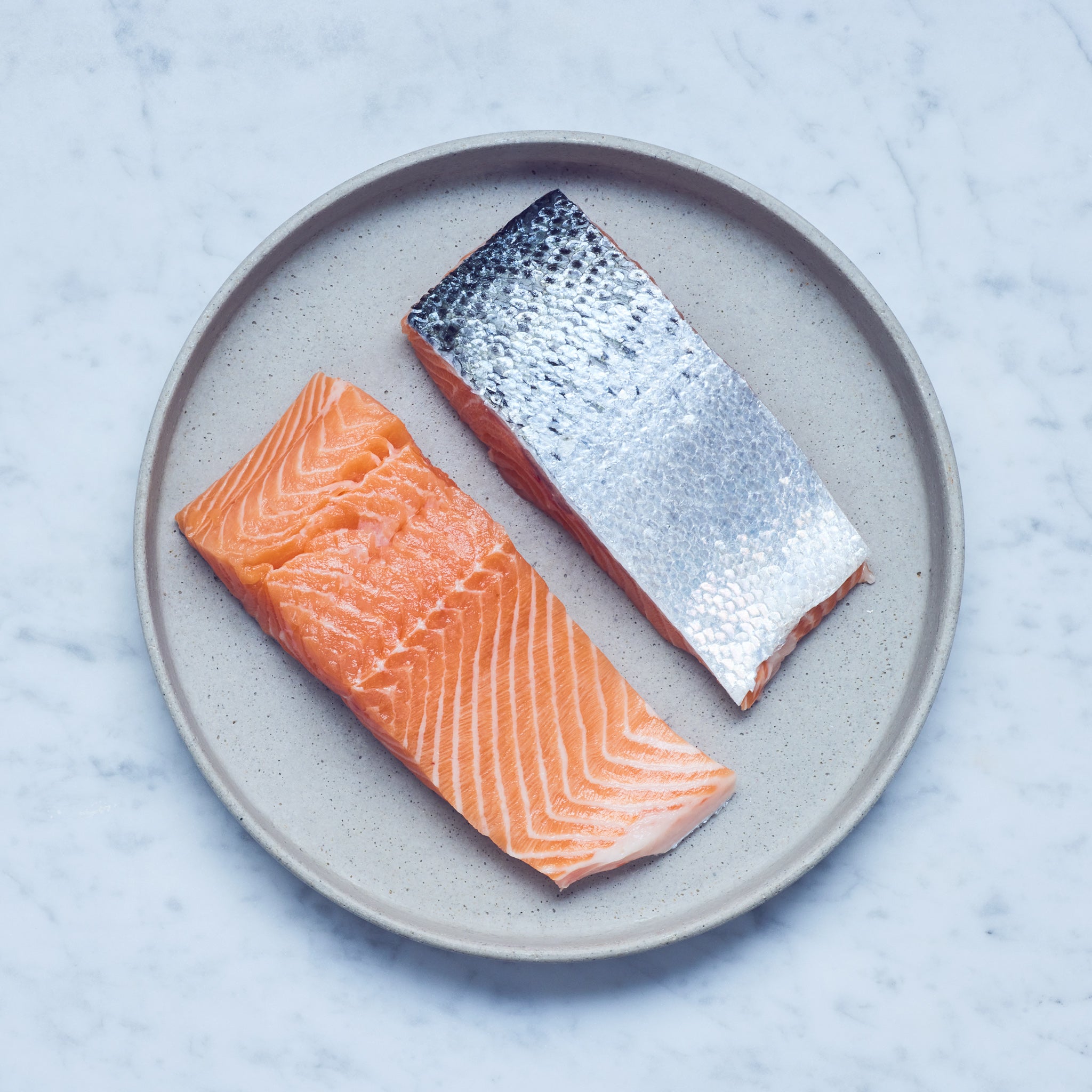 Lachsfilet kaufen. Ein runder Teller mit frischen Lachs-Filet in zwei Stücke geschnitten auf einer Marmoroberfläche.