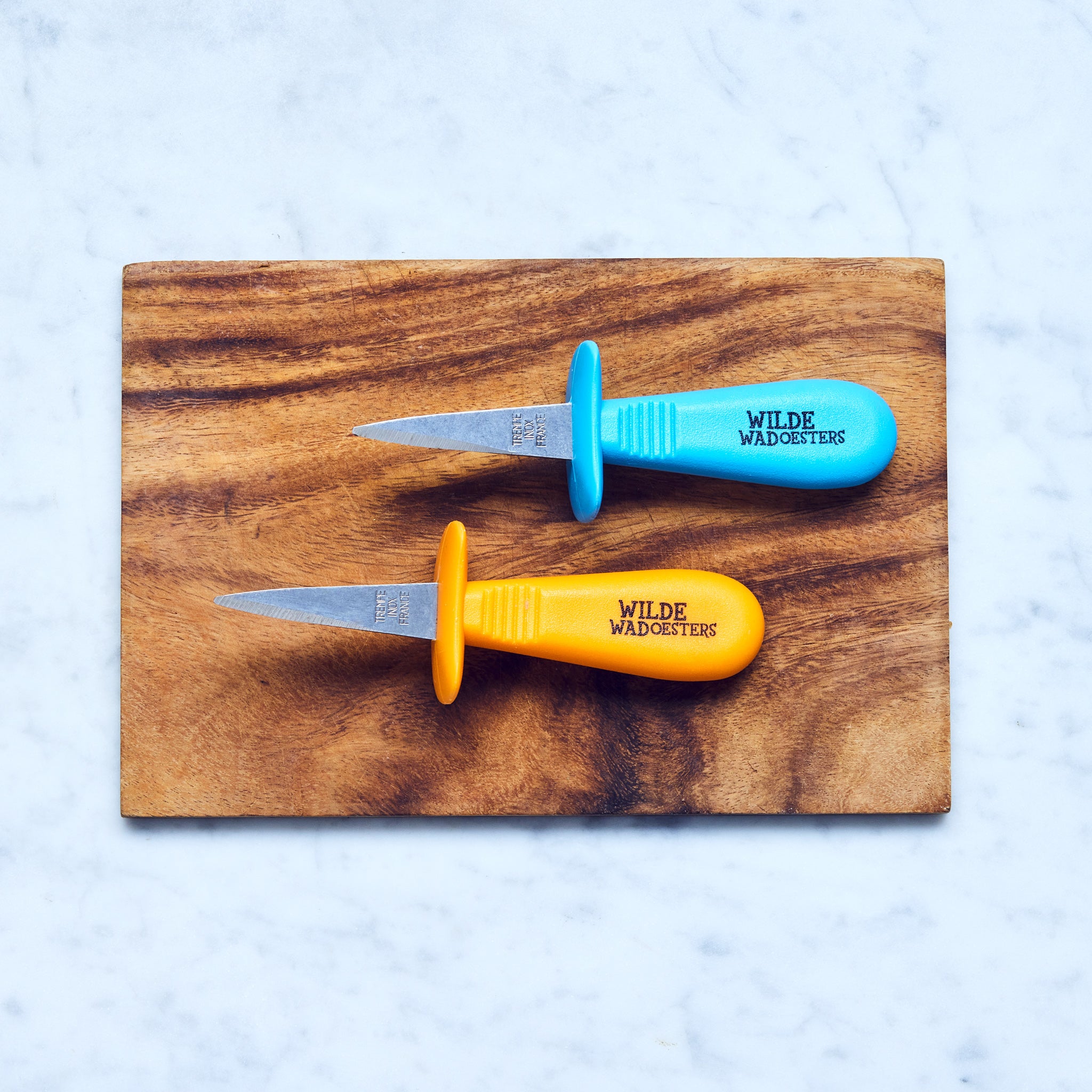 Zwei Austernmessern, blau und orange, auf einem Holzbrett.