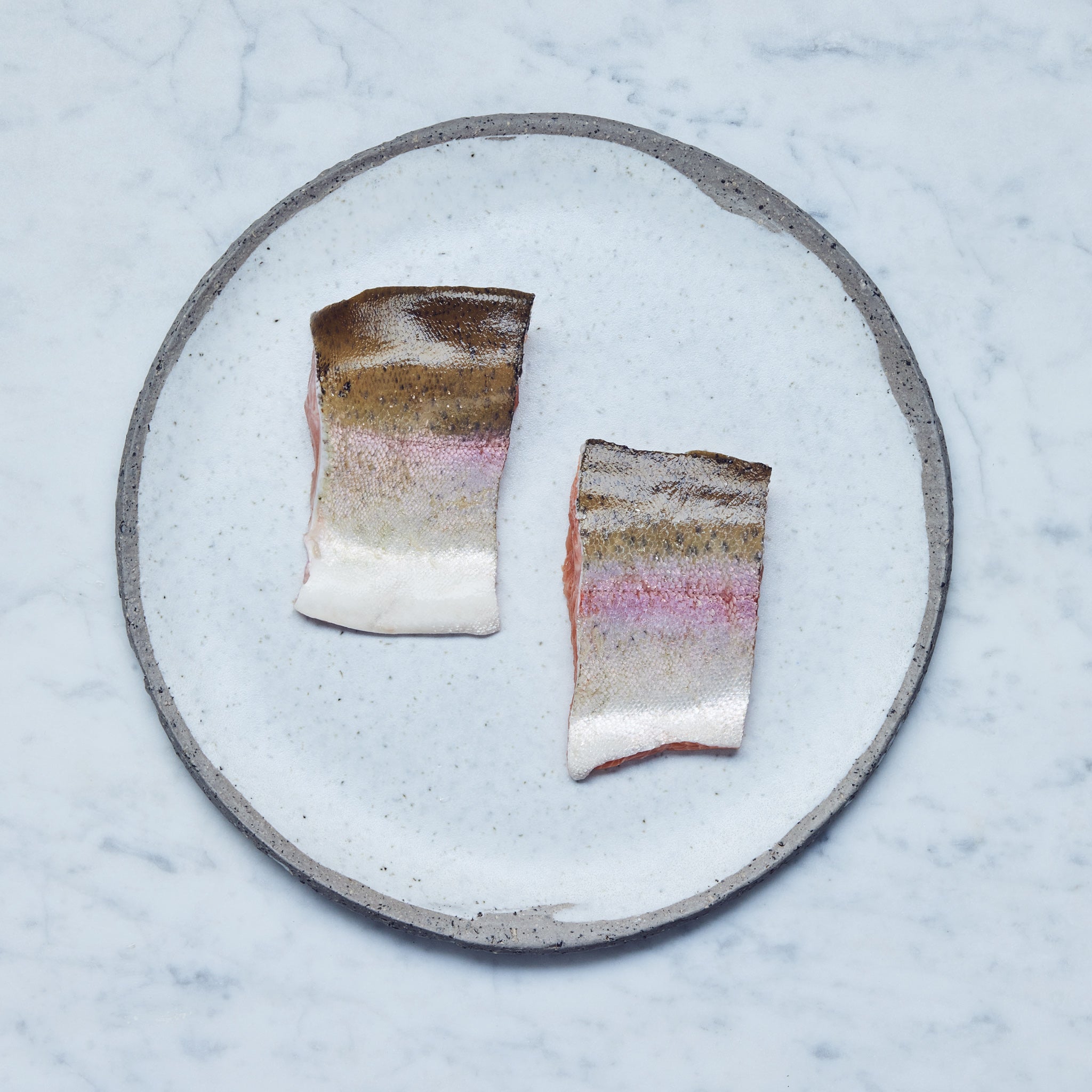 Ein runder Teller mit Bio-Lachsforellen-Filet in zwei Stücke geschnitten auf einer Marmoroberfläche.