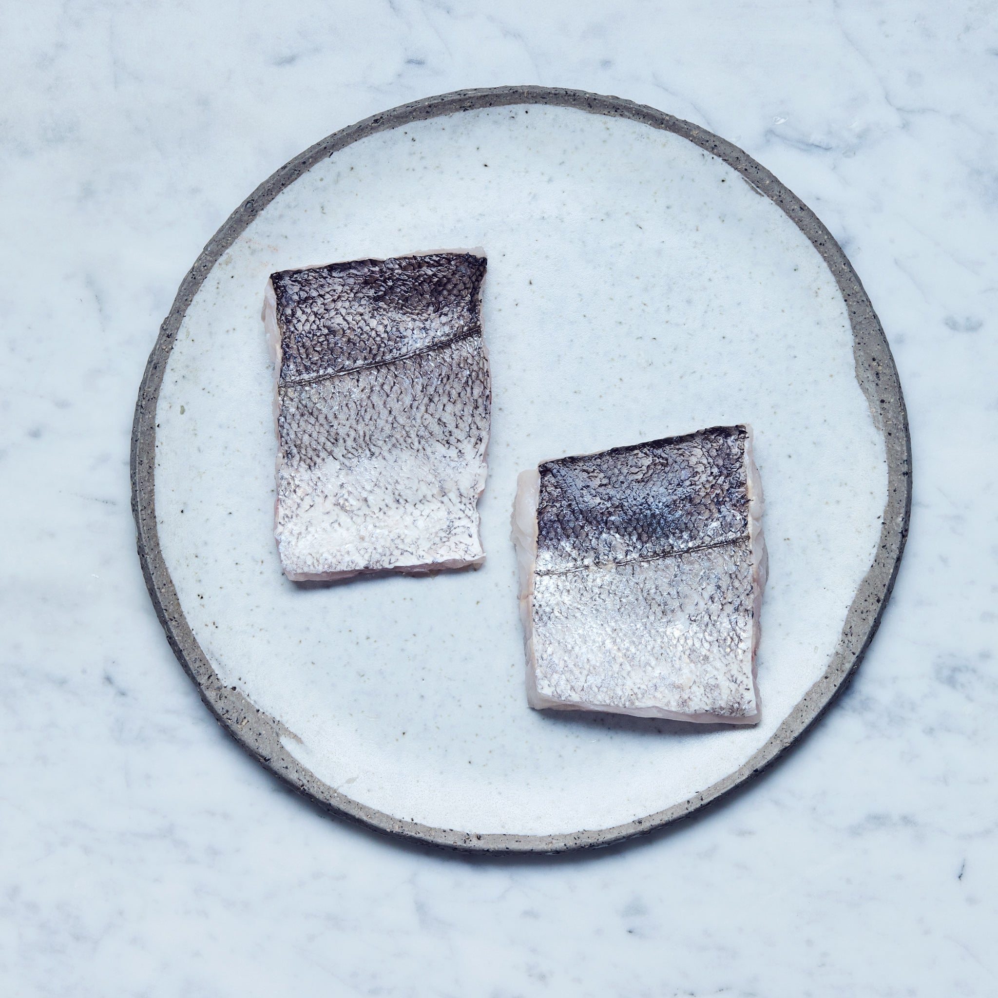Ein runder Teller mit frischen Seehecht-Filet in zwei Stücke geschnitten auf einer Marmoroberfläche.