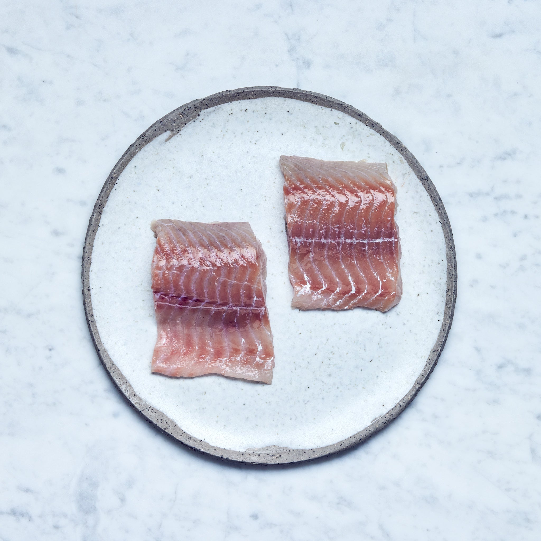 Ein runder Teller mit frischen Seelachs-Filet in zwei Stücke geschnitten auf einer Marmoroberfläche.