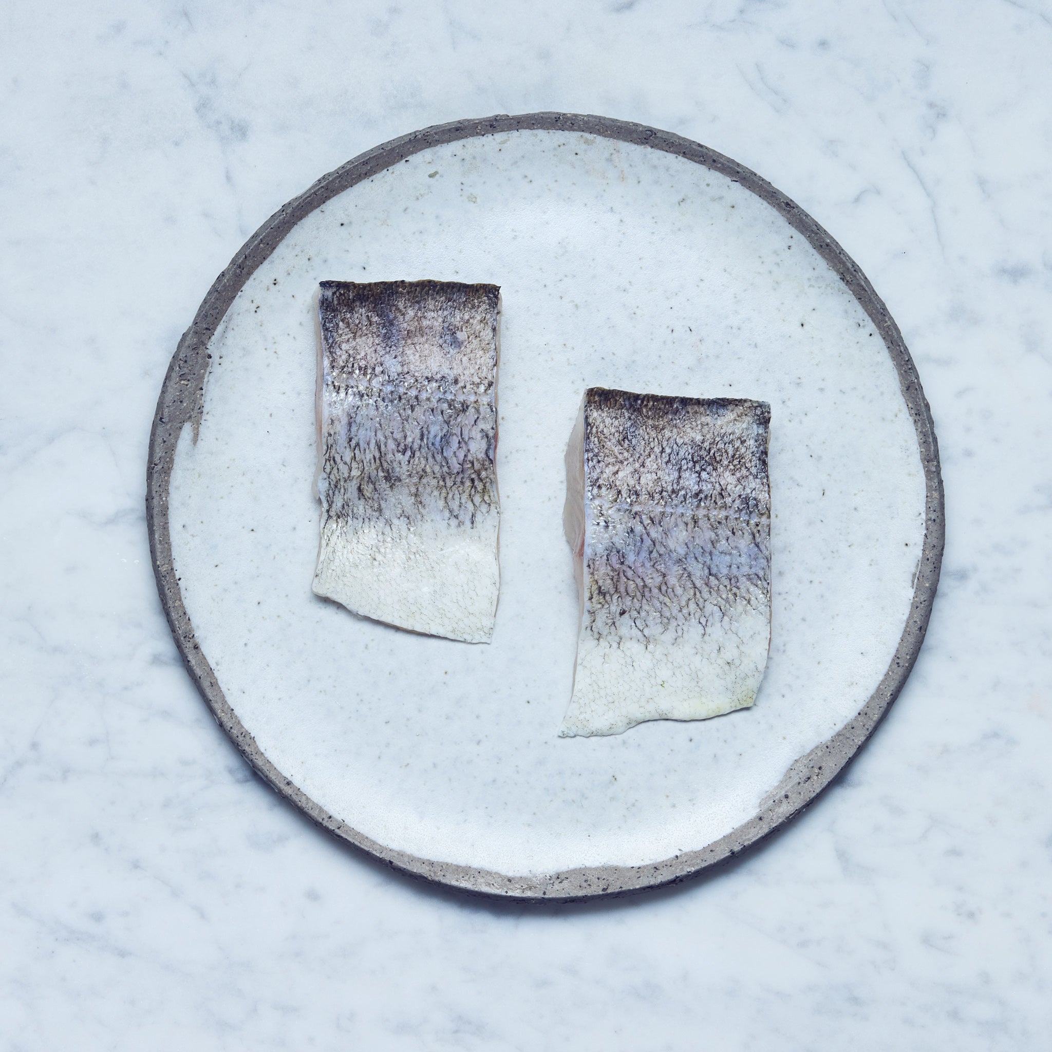 Ein runder Teller mit frischen Zander-Filet in zwei Stücke geschnitten auf einer Marmoroberfläche.